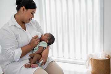Asian mother feeding milk from bottle milk to her 1-month-old baby black skin newborn son, is...