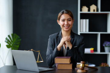 Beautiful Asian woman lawyer sitting at a table smiling happy with a laptop computer with law books hammer and scales hammer and scale. - obrazy, fototapety, plakaty
