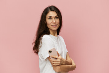 portrait of a beautiful, pleasant woman with long hair, in a light T-shirt holding a fashionable smartphone in her hands with her arms crossed on her chest. Horizontal photo on a pink background