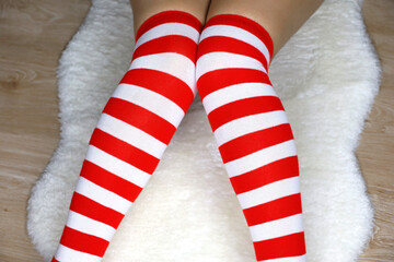 Fototapeta premium Legs of girl in Christmas knee socks sitting on white fur rug on wooden floor. Female outfit for New Year celebration