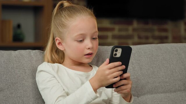 Beautiful pretty small girl pupil caucasian kid child daughter blogger sit on sofa at home use smartphone play mobile games chatting with classmates in social networks watching funny cartoons at home