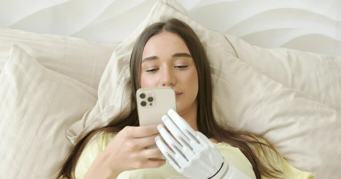 Girl with bionic arm using phone, reading social media and chatting in bed, woman with disability learning or working online remotely from home