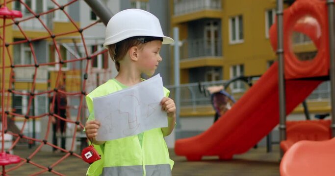 Excited preschooler imagines himself professional builder holding paper. Happy boy wearing white helmet looks around playing on kids playground