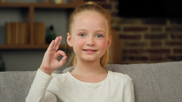 Portrait of caucasian happy child girl smiling kid sitting on sofa at room looking at camera showing ok gesture everything fine like sign positive good emotion. Little cute charming teen kid recommend