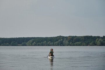 Obraz na płótnie Canvas Caucasian woman with dreadlocks sitting on board in lake and rowing with oar. Australian Shepherd in life jacket. Stand up paddle with pet. Concept animals actively spend time with owner.