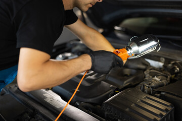The work of a car mechanic in a modern workshop, car service. The mechanic in blue overalls and safety glasses inspects the car while working with led lamp