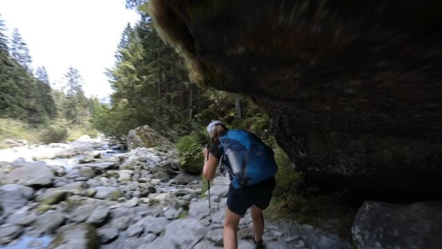Girl hiker walks on a path alongside a stream, passing under a rock banging his head. Adventure Backpacking in Val di Genova in the Adamello Brenta Park near the Nardis waterfalls, Trentino, Italy. 