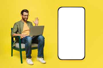 Positive man using laptop, sitting on chair near giant cellphone with empty white screen, having...