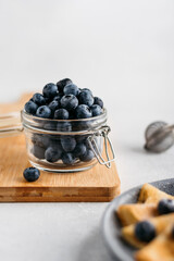 a jar of blueberries on a wooden board, next to pancakes. light background