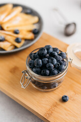 a jar of blueberries on a wooden board, next to pancakes. light background