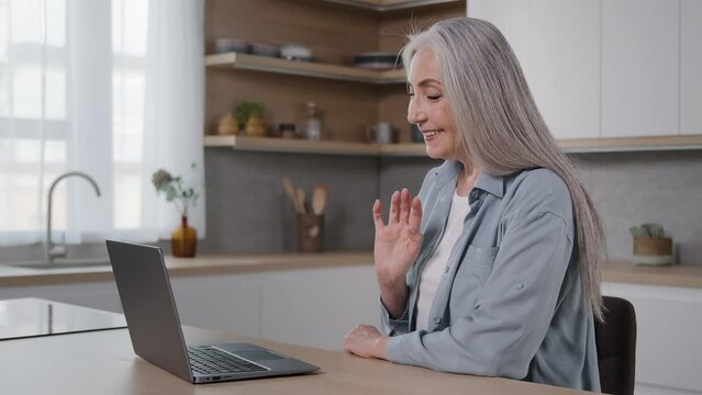 Caucasian senior woman elderly grey-haired lady sit in kitchen talk at computer webcam wave hand hello gesture video call conference chat with family distant conversation remote friendly communication