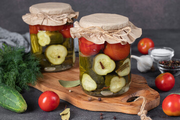 Pickled cucumbers with cherry tomatoes in two glass jars on brown background with fresh ingredients
