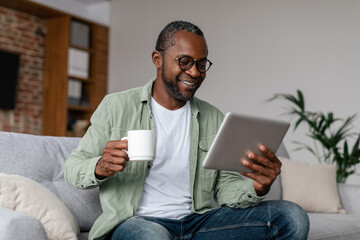 Smiling mature african american man in glasses drinking coffee from cup and watching video on tablet