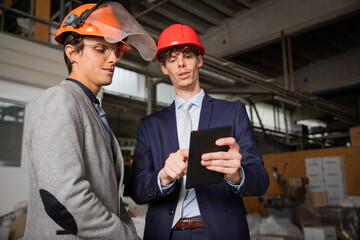 Two engineers discuss a project using a tablet while they are in the factory.