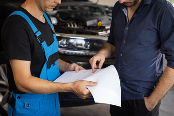 Specialist is showing info on tablet. Car service manager with a tablet, talking to mechanic man discussing car diagnostics and repairing. Mechanic talking to manager near vehicle in car service