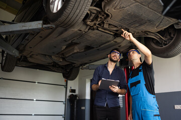 Fototapeta na wymiar Auto Service. Car Service Employees Inspect the Bottom and Skid Plates of the Car. Manager Checks Data on a Notebook and Explains the Breakdown to a Mechanic. Modern Workshop