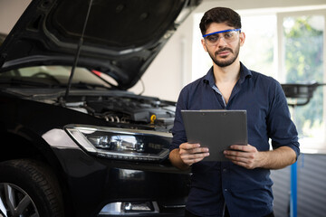 Portrait of caucasian bearded handsome man auto mechanic or manager in goggles smiling to camera in garage. Cheerful guy standing at cars service