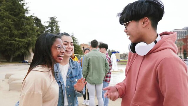 Group Of Three Multiethnic Teenage Students Standing Outside College Building tlking bout homeworks, at the background five friends laughing.