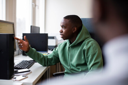 Male trainee in hooded shirt pointing at desktop screen while discussing in office