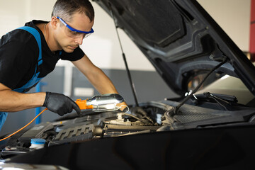 Caucasian Mechanic in Blue Overalls is Working on a Car in a Car Service. Repairman in Safety Glasses is Working on an Usual Car Maintenance. He Hangs Led Lamp. Modern Workshop