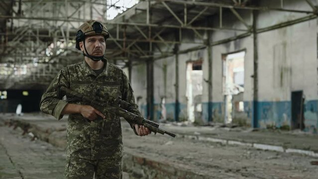 Strong military soldier with big rifle in his hands walking through abandoned old building. Member of armed forces guarding and patrolling destroyed steel factory.