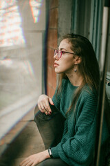 Refined intelligent woman in a green sweater and glasses looking out the window on an autumn day....