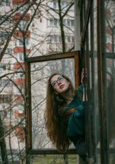 Attractive graceful girl with smart eyes and long hair looks out of the wooden window at the sky on a cold day. First autumn cold. A woman looks hopefully out of the window. Waiting.