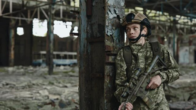 Caucasian military woman in uniform with weapon in hands hiding from enemies behind column on destroyed steel plant. Protective mission during war.