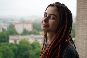 Smiling pretty woman with red dreadlocks in a gray sweater looks at the city from the balcony after the rain. Sensual girl on the balcony. Autumn mood. 