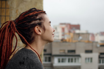 Young woman with red dreadlocks in a gray sweater looks at the city from the balcony after the rain. Sensual girl on the balcony. Autumn mood. Drops of water on a woman's skin. Fresh air.