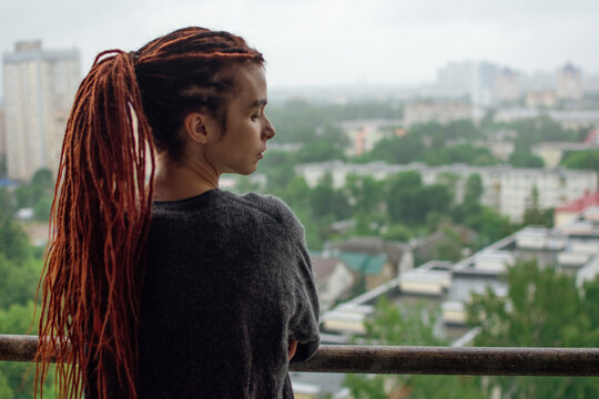 Young girl with red dreadlocks in a gray sweater looks at the city from the balcony after the rain. Sensual girl on the balcony. Autumn mood. Fresh air. 