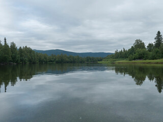 View of wide calm Tarra river from boat at Kvikkjokk, with forest and grass shore and green hills. Northern landscape in Swedish Lapland. End of Padjelantaleden hiking trail. Cloudy summer day