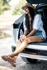 Beutiful woman sitting in the trunk of the car while drinking water in the forest.