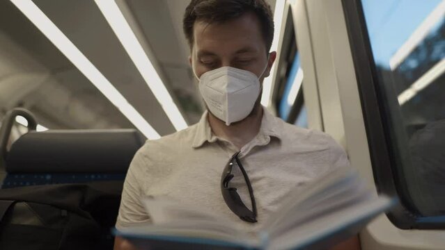 Male passenger in mask sits in passenger seat on modern train in Germany and reads book during coronavirus pandemic. Male in protective mask reading a book on a railroad trip sitting by the window 