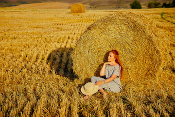 Naklejka na ściany i meble A beautiful young woman in a hat and a summer dress is sitting on a sheaf of hay in a field. Rural nature, wheat field, rest in the country, unity with nature