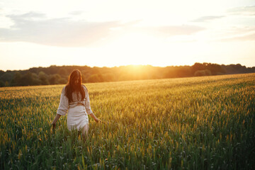 Woman walking in wheat field in warm sunset light. Stylish young female in rustic dress holding...