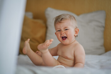 happy cute naked one year old happy blond baby boy sitting at home on a cozy bed after bathing and smiling or flirting into the Camera 