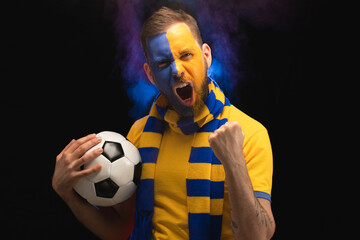 Portrait of euphoric soccer supporter man with yellow-blue painted face is making winners gesture clenching his fist and posing with a ball in hand over black background with smoke effect - 527436510