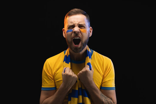 Portrait of excited young soccer supportred man in yellow-blue t-shirt and scarf and painted face, cheering for his favourite team