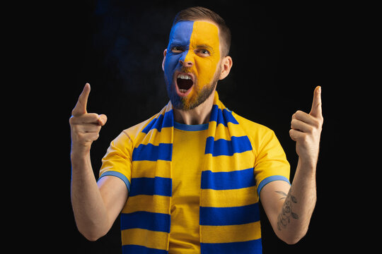Portrait of excited shouting soccer supporter man in yellow-blue t-shirt and scarf and with painted face, cheering for his favourite team, isolated over black background with smoke effect