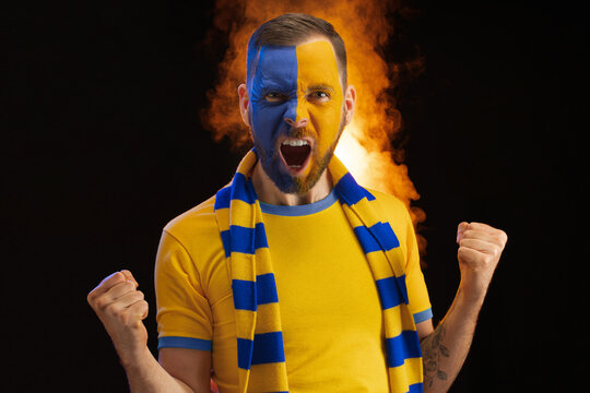 Portrait of happy excited sport supporter man with yellow-blue painted face, celebrating his team victory, clenching fists and screaming yes, with fire and smoke effects on the dark background