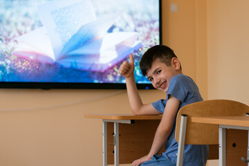 Elementary school student sits on the bench at the school desk, makes the good sign with his hand, on the background of the screen, back to school