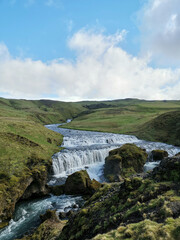 River going through mountains and green grass with water flowing stone waterfall with blue sky and white cloud vertical idyllic nature