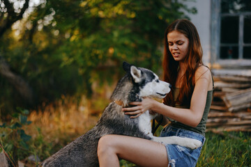 Obraz premium A young woman plays with her husky dog ​​in nature near the house in the forest against the backdrop of the setting sun on an autumn evening