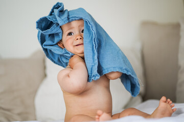 Fototapeta na wymiar mega cute naked almost one year old blond baby boy sitting & laughing at home on a cozy bed after bathing and playing with a blue muslin fabric burp cloth while making nonsense jokes