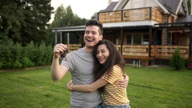 Very Happy man and asian woman is owners shake key and hug each other in front of their newly bought home. Laugh and smile. Young married couple or family buy a new house. Excited clients purchasing