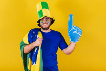 Brazilian caucasian man, soccer fan from Brazil, wearing hat and foam finger, cheering and smiling.
