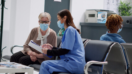 Asian nurse consulting senior woman with face mask in hospital reception lobby, talking about disease diagnosis and healthcare. Specialist giving advice about covid 19 prevention. Handheld shot.