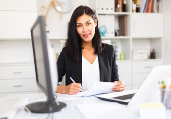 Smiling woman working with papers and laptop in office - obrazy, fototapety, plakaty