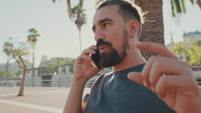 Young man with beard sits on bench emotionally talking on cellphone. Backlight